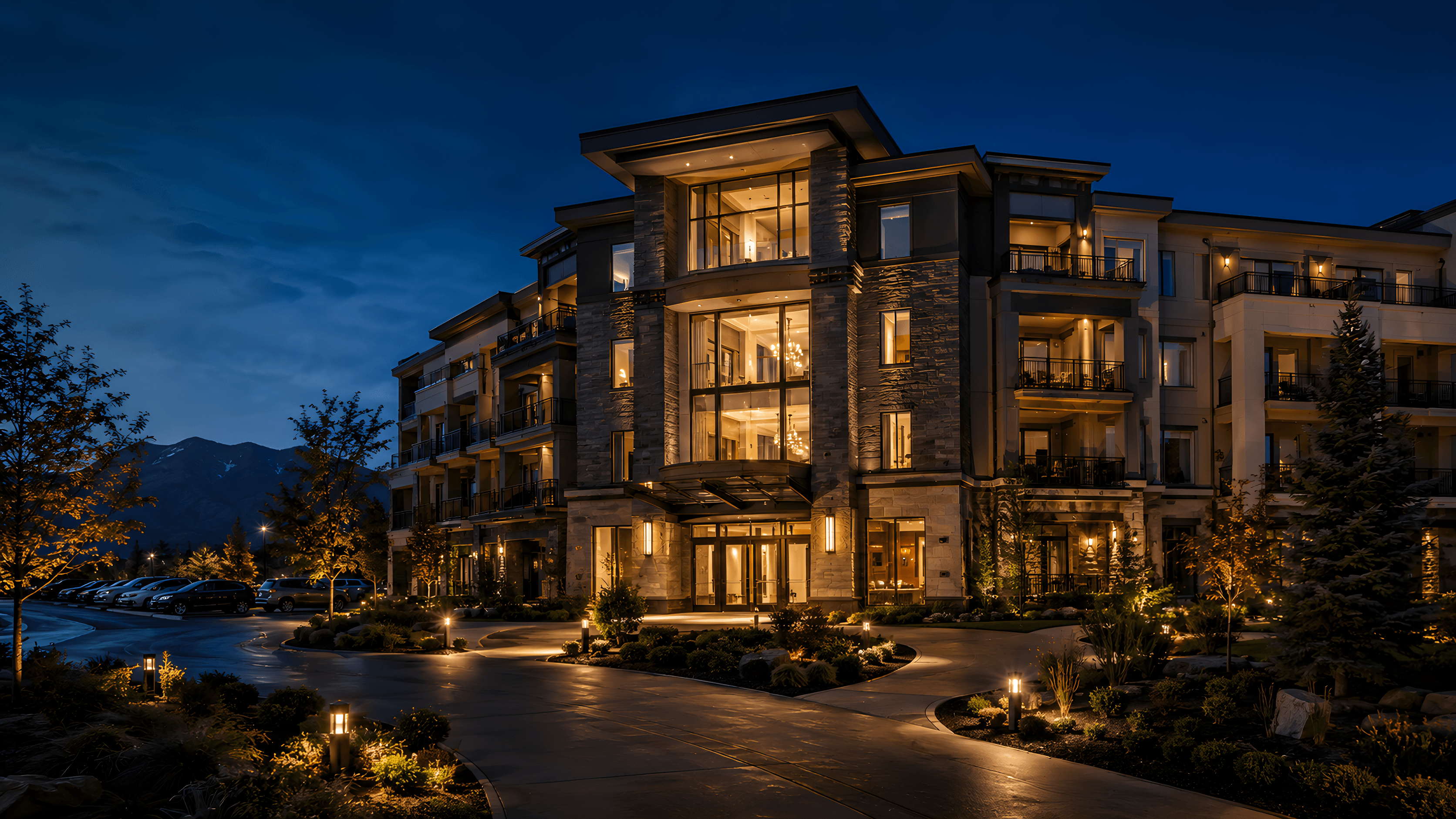 Modern multi-family apartment community at dusk with warm-lit balconies and a landscaped entry drive